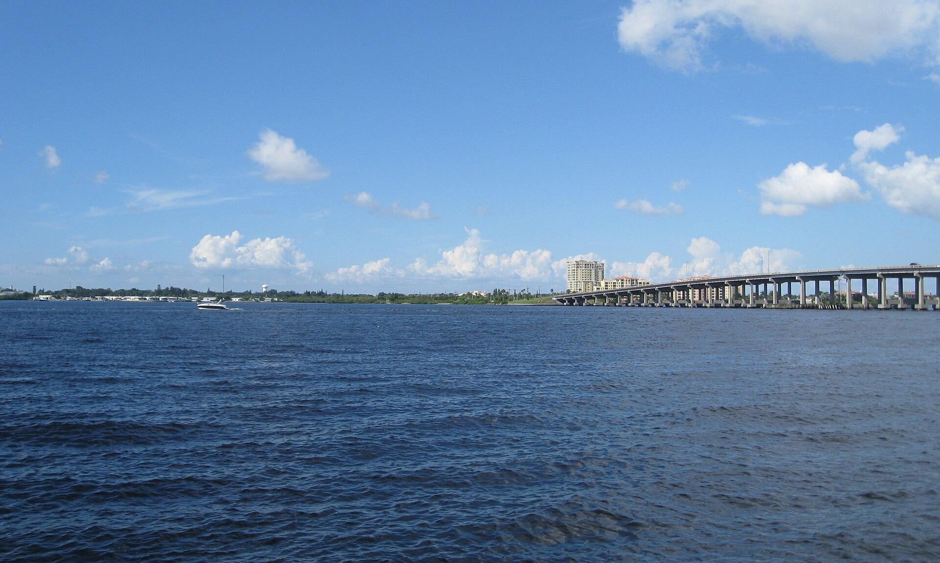 Manatee River waterfront and DeSoto Bridge viewed from the Bradenton Riverwalk, with blue water, passing boat, and the downtown Bradenton skyline under a sunny Florida sky